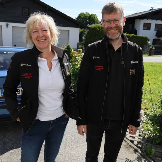 Eine Frau und ein Mann stehen lächelnd für ein Foto draußen. Sie tragen beide schwarze Jacken mit Logos und stehen vor einem Haus mit Garage.
