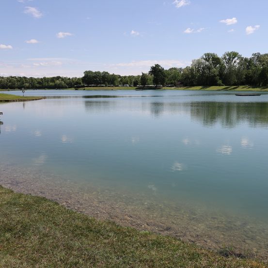 Ein ruhiger See mit klarem Wasser, das den blauen Himmel widerspiegelt, umgeben von üppigem grünen Bäumen und Gras. Am Ufer befindet sich ein kleiner Rasenbereich.