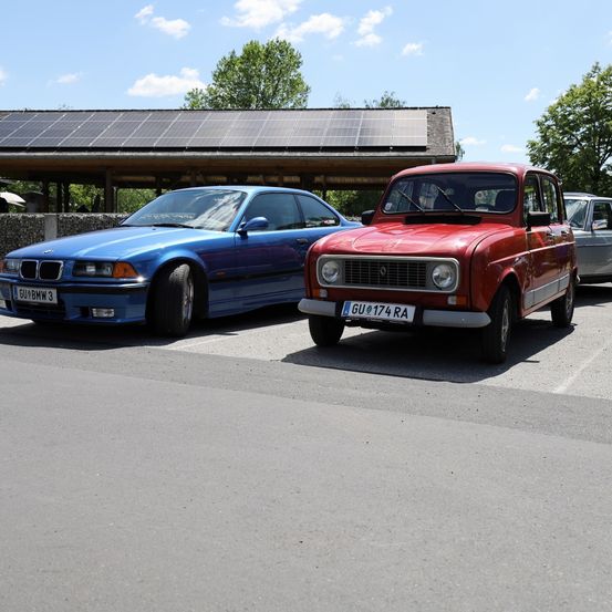 Drei Autos sind auf einem Parkplatz unter einem Gebäude mit Solarpanelen geparkt. Ein blauer BMW und ein roter Renault sind nebeneinander geparkt.