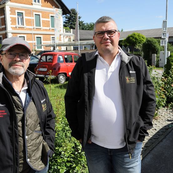 Zwei Männer stehen vor einem Gebäude, einer mit einer Brille und einem Hut, der andere in einem weißen Shirt und einer schwarzen Jacke. Hinter ihnen sind ein rotes Auto und ein Gebäude zu sehen.