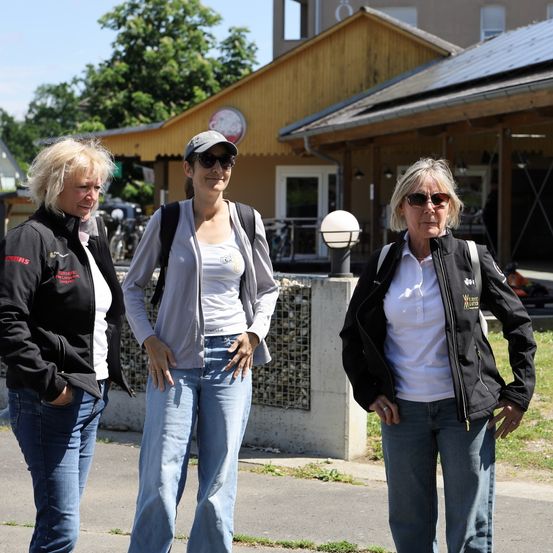 Drei Frauen in Jacken stehen vor einem gelben Gebäude mit Solarmodulen. Die Frau in der Mitte trägt eine Mütze und eine Sonnenbrille, während die anderen ihre Hände in den Taschen haben.