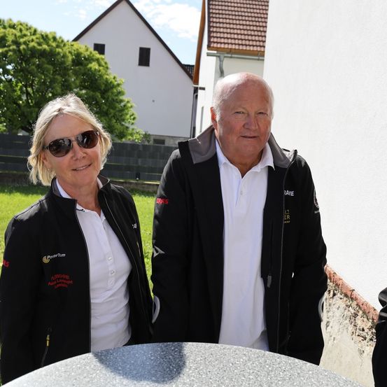Ein Mann und eine Frau stehen lächelnd vor einem Haus mit rotem Dach. Sie tragen passende schwarze Jacken mit roten Logos. Die Frau trägt eine Sonnenbrille und der Mann ein weißes Hemd. Dahinter sind eine Wand und ein Baum sichtbar.