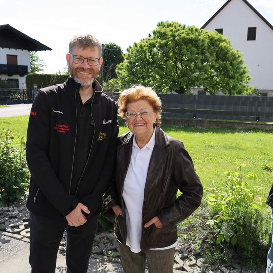 Ein Mann und eine Frau stehen zusammen vor einem Haus mit Zaun. Der Mann trägt einen schwarzen Mantel mit roten Buchstaben, und die Frau trägt eine Lederjacke und eine Brille. Sie lächeln.
