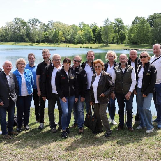 Eine Gruppe von Menschen steht vor einem See, einige tragen Sonnenbrillen und Jacken. Im Hintergrund befinden sich Bäume und ein Rasen.
