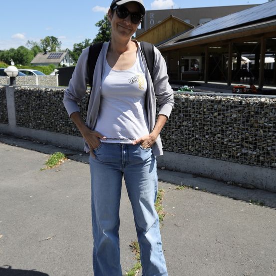 Eine Frau in einem weißen Tanktop und Sonnenbrille posiert vor einer Steinmauer, mit einem Gebäude mit Solarpanelen im Hintergrund.