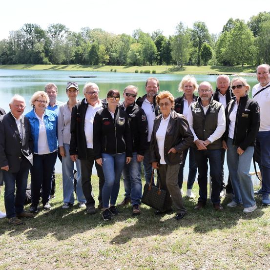 Eine Gruppe von Menschen steht vor einem See, in Freizeitkleidung gekleidet. Sie scheinen an einem sonnigen Tag für ein Foto zu posieren.