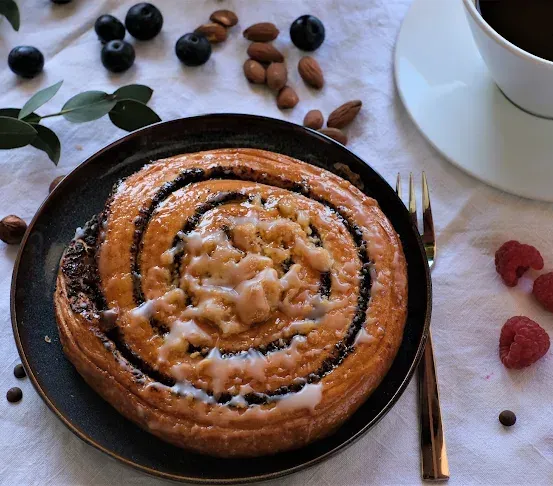 Ein Spiralgebäck mit schwarzer Füllung und weißer Glasur auf einem schwarzen Teller, umgeben von Nüssen, Beeren und einer Tasse Kaffee.