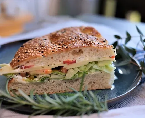 Ein halbes Sandwich mit Sesamkörnern auf einem Teller, mit Rosmarin garniert und von Blättern umgeben.