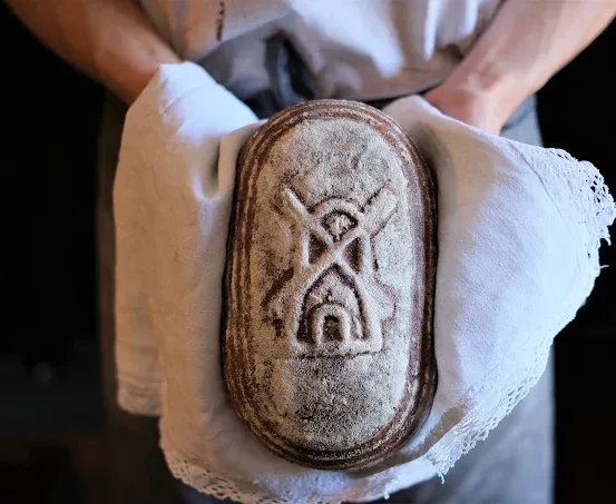 Eine Person mit weißen Handschuhen hält ein rustikales, rechteckiges Brot mit einem Kreuz- und Hausdesign darauf.