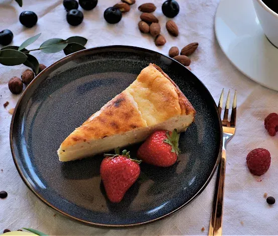 Ein Stück Käsekuchen auf einem dunklen Teller mit zwei Erdbeeren und einer Gabel, umgeben von Beeren, Nüssen und einer Tasse Kaffee.