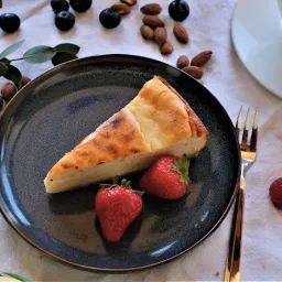 Ein Stück Käsekuchen auf einem dunklen Teller mit zwei Erdbeeren und einer Gabel, umgeben von Beeren, Nüssen und einer Tasse Kaffee.