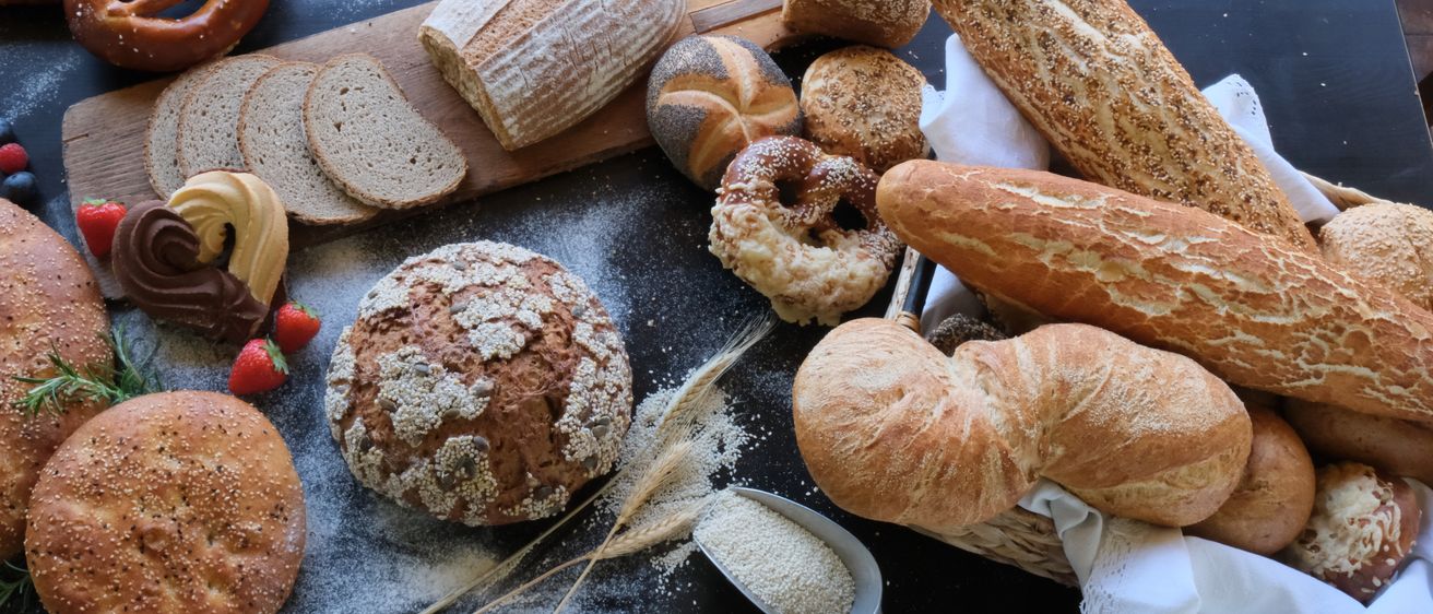 Auf einem Tisch sind verschiedene Brotsorten zu sehen, darunter ein Laib, Brezeln und Donuts, mit verstreuten Körnern und einer Schaufel Mehl.