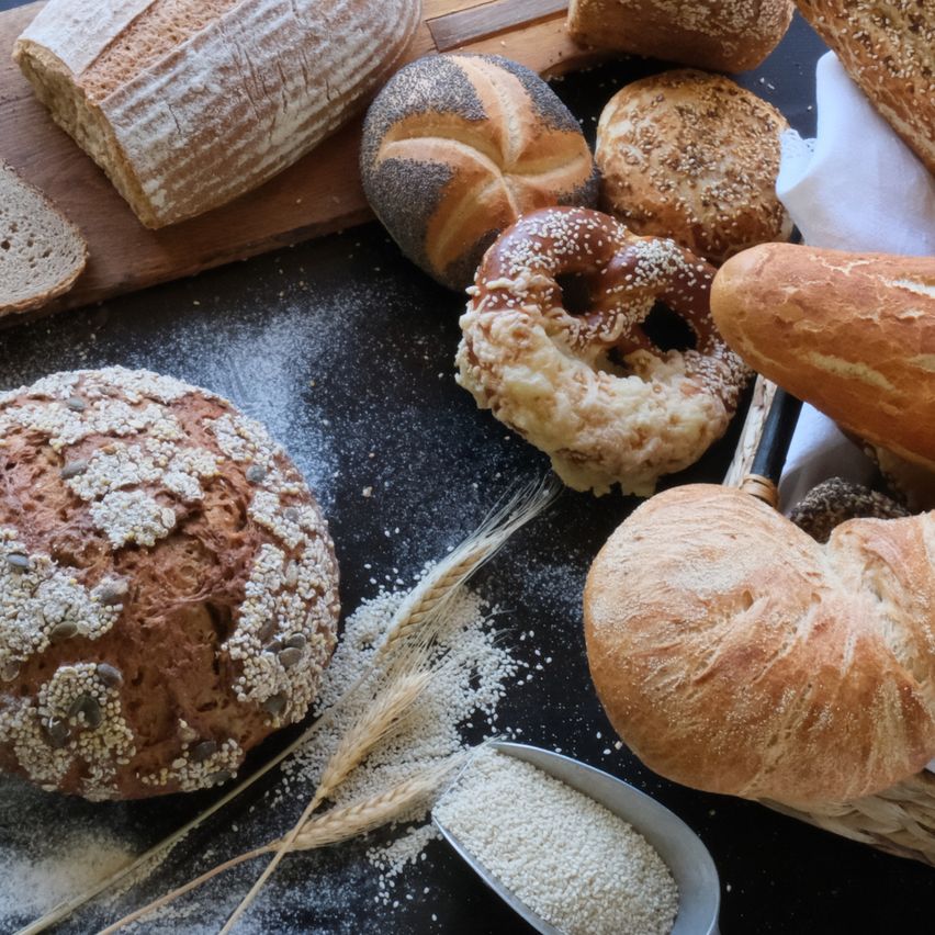 Auf einem Tisch sind verschiedene Brotsorten zu sehen, darunter ein Laib, Brezeln und Donuts, mit verstreuten Körnern und einer Schaufel Mehl.
