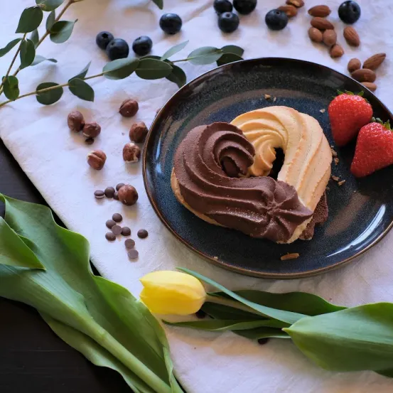 Ein herzförmiges Gebäck mit Schokolade und Sahne auf einem Teller, umgeben von Erdbeeren, Blaubeeren, Haselnüssen und Mandeln, mit grünen Blättern und einer gelben Tulpe.