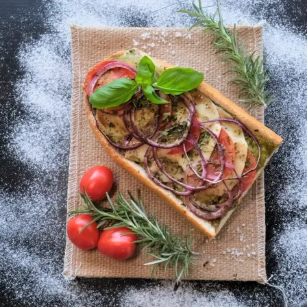 Ein Stück Brot mit roten Zwiebeln und Basilikum darauf, mit Kirschtomaten und Rosmarin an der Seite.