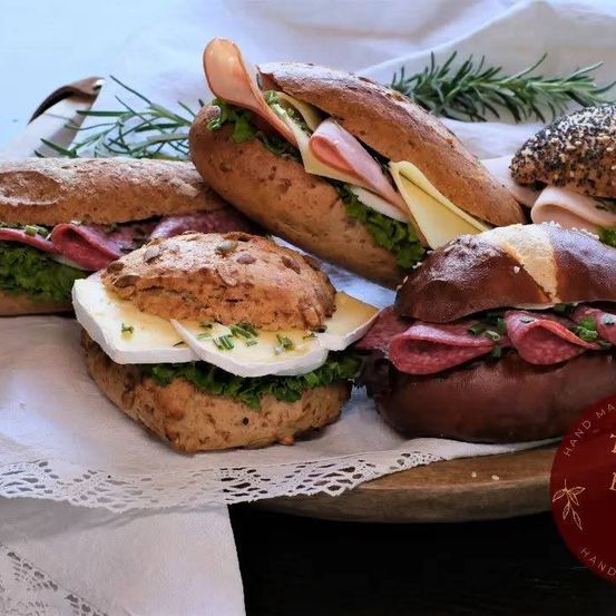 Ein Holzbrett zeigt verschiedene Sandwiches, darunter eines mit Käse, Salat und Schinken, ein weiteres mit einer Mischung aus Fleisch und ein drittes mit einer Sesambrötchen.