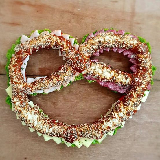Zwei Brezeln sind in Herzform auf einer Holzoberfläche angeordnet, gefüllt mit Käse, Salami und mit grünen Kräutern garniert.