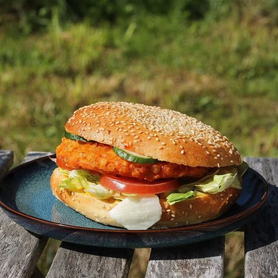 Ein Hühnchensandwich mit Salat, Tomate und Gurke auf einem Sesambrötchen, serviert auf einem blauen Teller auf einem Holztisch mit einem unscharfen Grashintergrund.