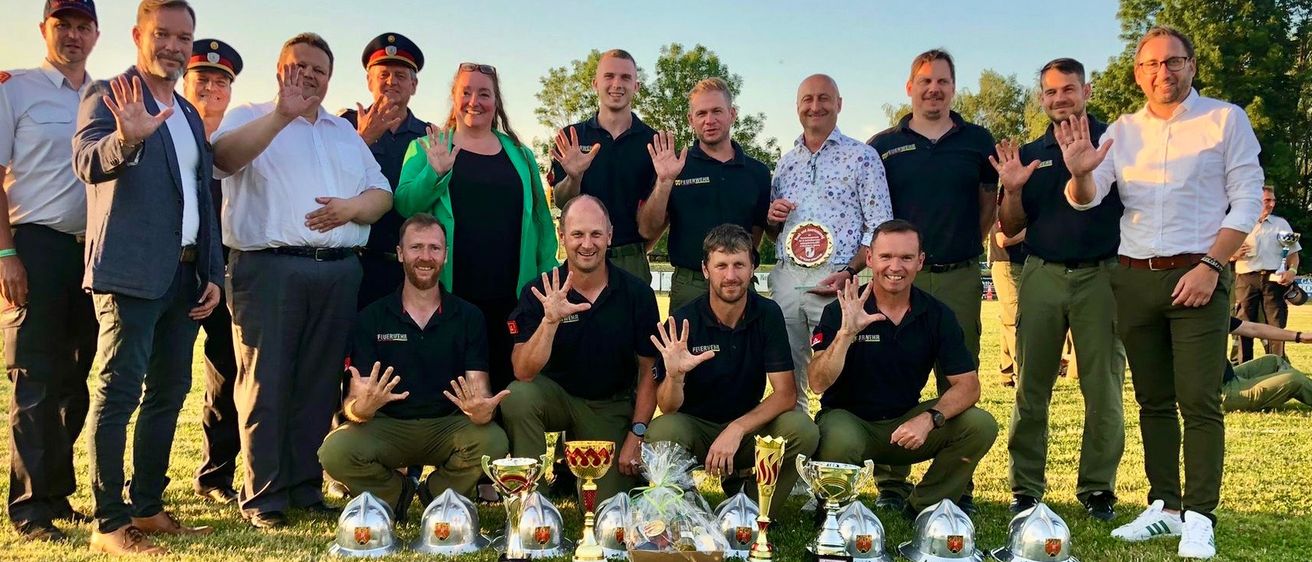 Eine Gruppe von Menschen posiert für ein Foto auf einem Rasenfeld, hält Trophäen und lächelt. Einige tragen Hüte, Gürtel und Uhren. Sie haben ein Abzeichen auf ihren Hemden.