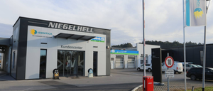 Die Vorderseite eines modernen Gebäudes mit einem Schild, das Niegelhell liest, mit einem Kundencenter-Eingang, großen Glastüren und einem weißen Auto, das in der Nähe geparkt ist.