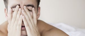 Ein Mann liegt auf einem Bett mit geschlossenen Augen und bedecktem Gesicht, sieht erschöpft aus.