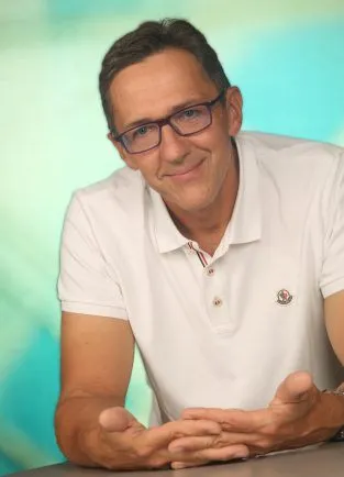 Ein Mann mit Brille, der ein weißes Poloshirt trägt, lächelt und sitzt mit zusammengepressten Händen an einem Tisch.