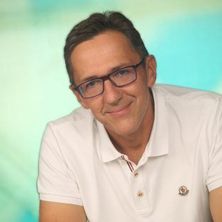 Ein Mann mit Brille und einem weißen Poloshirt mit einem Logo auf der linken Brust sitzt lächelnd am Tisch mit zusammengefalteten Händen.