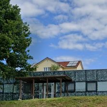 Ein Gebäude mit Solarpanels auf dem Dach, marmortexturierten Wänden und einer Holzveranda steht auf einem Grashügel unter einem blauen Himmel mit Wolken.
