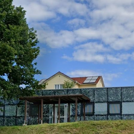 Ein Gebäude mit Solarpanels auf dem Dach, marmortexturierten Wänden und einer Holzveranda steht auf einem Grashügel unter einem blauen Himmel mit Wolken.