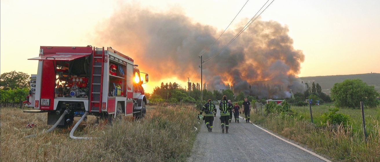 Feuerwehrleute in Uniform gehen einen Weg hinunter zu einem Feuerwehrwagen. Im Hintergrund steigt Rauch auf.