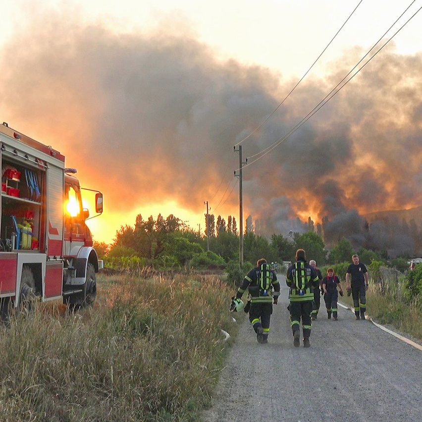 Feuerwehrleute in Uniform gehen einen Weg hinunter zu einem Feuerwehrwagen. Im Hintergrund steigt Rauch auf.