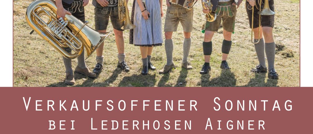 Eine Gruppe von Männern und Frauen in traditioneller bayerischer Kleidung steht mit Musikinstrumenten auf einem Feld. Der Mann auf der linken Seite spielt Tuba, während die anderen Trompeten, ein Akkordeon und ein anderes Instrument spielen. Sie stehen vor einem Berg.