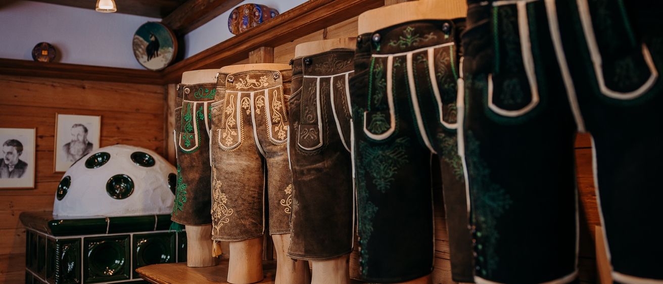 Ein Holzregal mit einer Reihe von Mannequins, die traditionelle bayerische Lederhosen tragen. Jedes Paar hat unterschiedliche Stickereien. Oben befindet sich eine Deckenleuchte und dekorative Gegenstände.