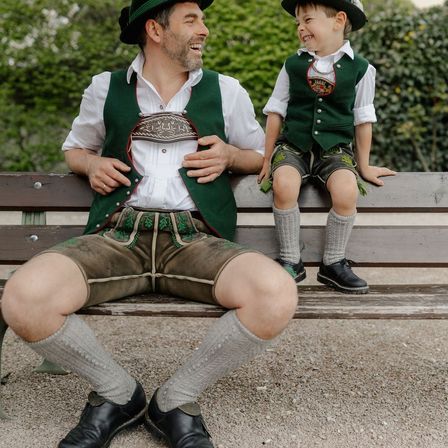 Ein Mann und ein Kind in traditioneller bayerischer Kleidung sitzen auf einer Bank und lächeln. Das Kind trägt eine grüne Weste mit Logo, Shorts und Socken. Der Mann trägt eine grüne Weste, Hose und Socken.