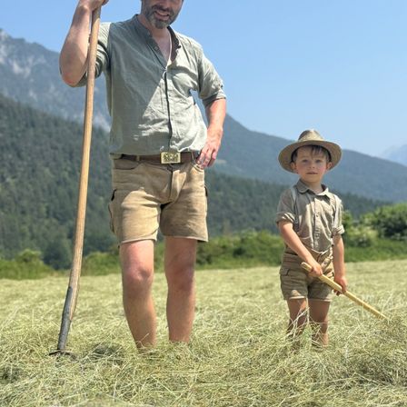 Ein Mann und ein kleiner Junge stehen in einem Feld, der Junge hält eine Harke. Der Mann hält einen langen Holzstab. Sie tragen beide Shorts und Hemden. Im Hintergrund sind Berge.