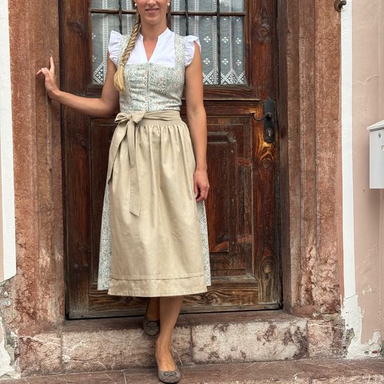 Eine Frau in einem traditionellen Kleid mit geflochtenen Haaren steht auf den Stufen vor einer Holztür mit einem Fenster.