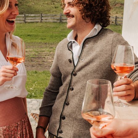 Ein Mann und eine Frau lächeln, jeder hält ein Glas mit Roséwein, stehend in der Nähe eines Grasfeldes mit einem Holzzaun im Hintergrund.