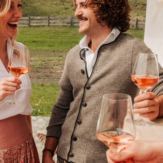 Ein Mann und eine Frau lächeln, jeder hält ein Glas mit Roséwein, stehend in der Nähe eines Grasfeldes mit einem Holzzaun im Hintergrund.