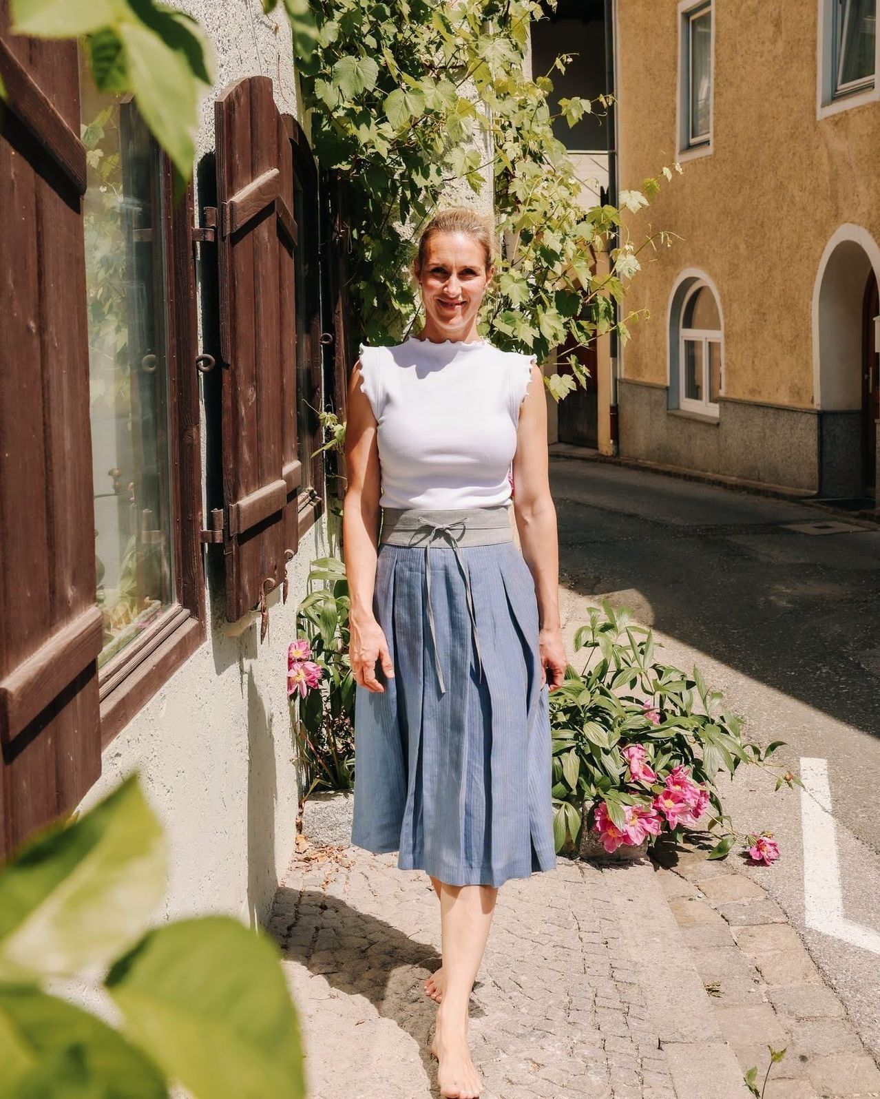 Eine Frau steht auf einer Kopfsteinpflasterstraße, lächelt und trägt ein weißes Oberteil und einen blauen Rock. Hinter ihr befindet sich ein Gebäude mit Bogenfenstern und einem geschlossenen Fensterladen.