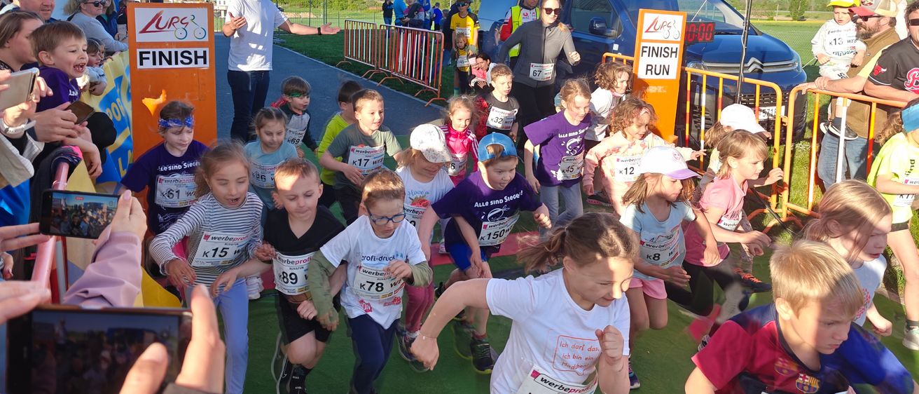 Eine Gruppe von Kindern nimmt an einem Laufevent teil, alle tragen Startnummern auf ihren Shirts. Einige tragen Mützen, Brillen und Turnschuhe. Zuschauer und ein Van sind im Hintergrund zu sehen.