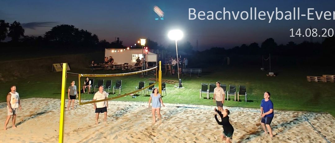 Ein Beachvolleyballspiel bei Nacht mit einigen spielenden Personen auf dem Sand. Eine Frau in Blau springt, während andere stehen und zuschauen. Straßenlaternen und ein Zelt sind im Hintergrund.