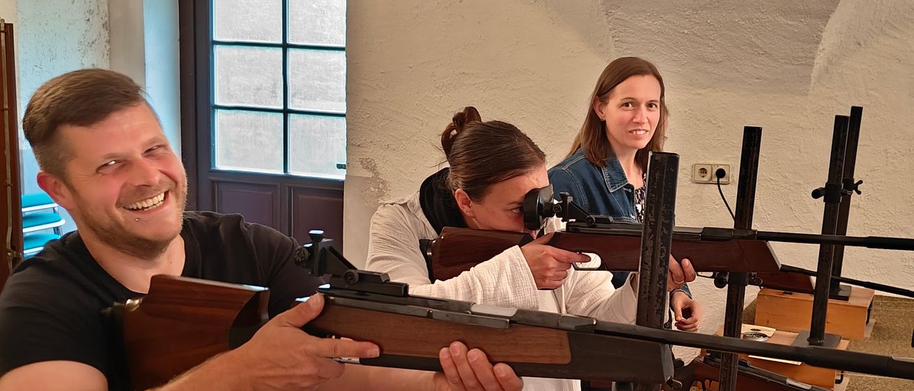 Eine Frau zielt mit einem Gewehr im Innenraum, wobei eine andere Frau hinter ihr steht. Ein Fenster und eine Holzwand sind im Hintergrund zu sehen.