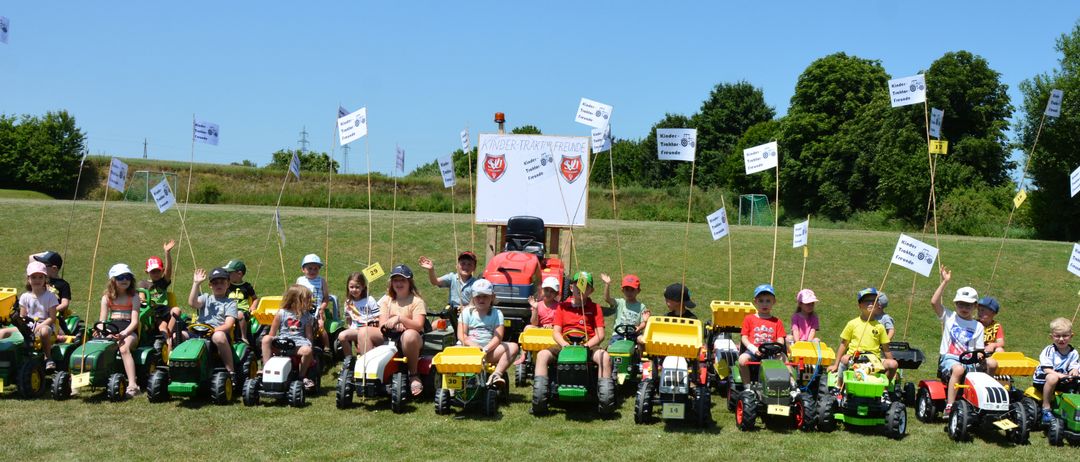 Kinder sitzen auf Spielzeugtraktoren auf einer Wiese. Ein weißes Schild mit dem Text Kinder-Traktor-Freunde ist auf einem Ständer in der Mitte montiert.