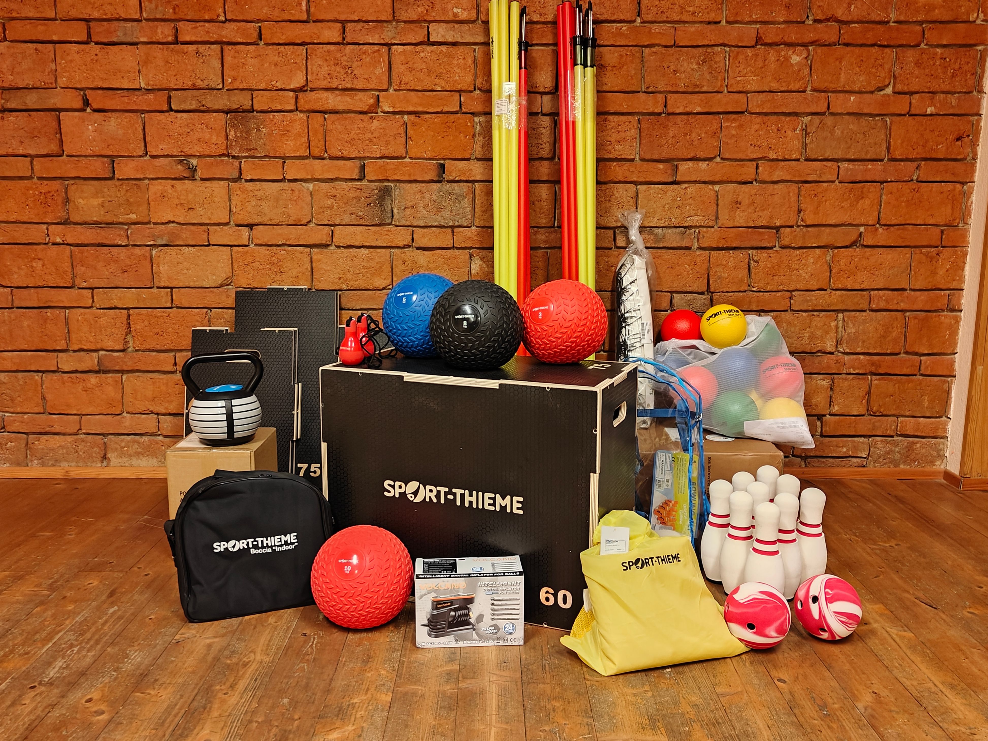Verschiedene Sportgeräte auf einem Holzboden ausgestellt, darunter eine schwarze Box mit der Aufschrift Sport-Thieme, eine Kettlebell, Bowlingkegel und mehrere farbige Bälle.