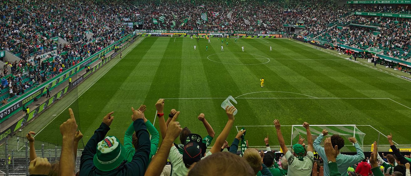 Ein Fußballspiel im Weststadion, Fans jubeln. Die Anzeigetafel zeigt 3-1. Spieler in grünen und gelben Trikots sind auf dem Feld.