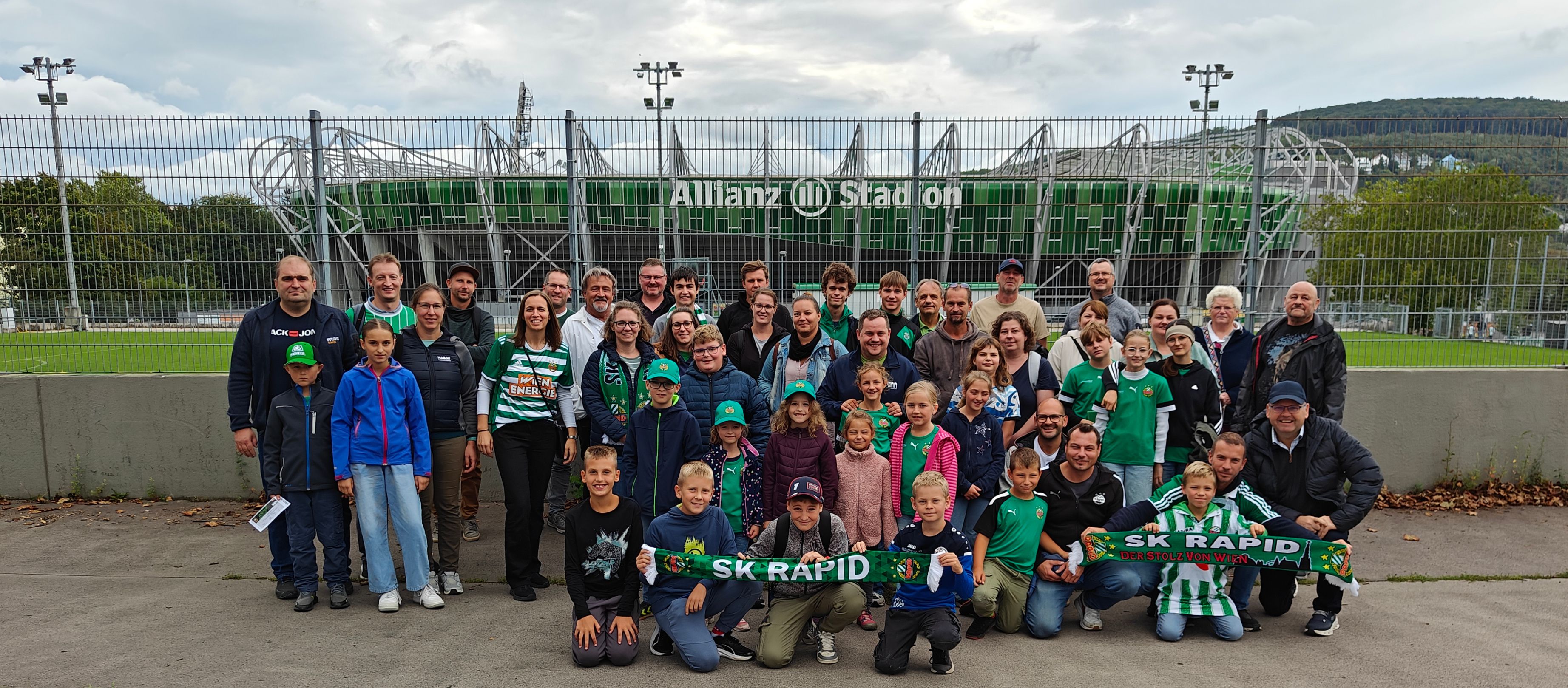Eine Gruppe von Menschen, wahrscheinlich Fans, posiert für ein Foto vor dem Allianz-Stadion. Sie tragen verschiedene Team-Trikots und halten ein Banner mit den Worten SK Rapid.