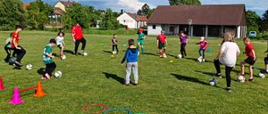 Mehrere Kinder in verschiedenen Uniformen spielen auf einem Rasenfeld Fußball mit mehreren farbigen Reifen und Bällen um sie herum. Ein Mann in einem roten Mantel und Turnschuhen geht im Vordergrund.