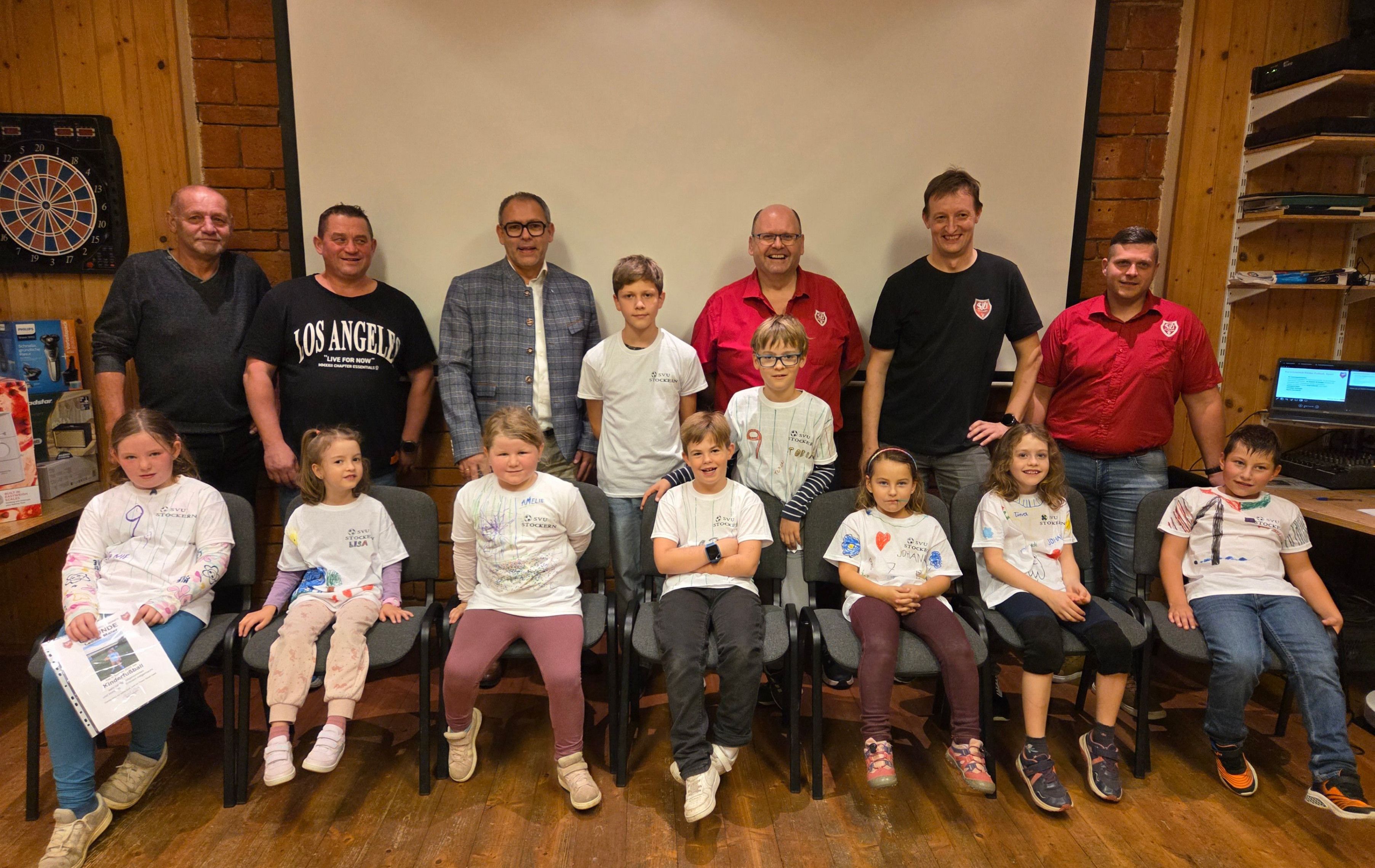 Eine Gruppe von Erwachsenen und Kindern posiert für ein Bild vor einer weißen Wand, alle tragen weiße T-Shirts mit farbenfrohen Designs. Die Kinder sitzen auf Stühlen, und die Erwachsenen stehen hinter ihnen. Das Bild zeigt einen Moment der Einheit und des Engagements in einer Lernumgebung.