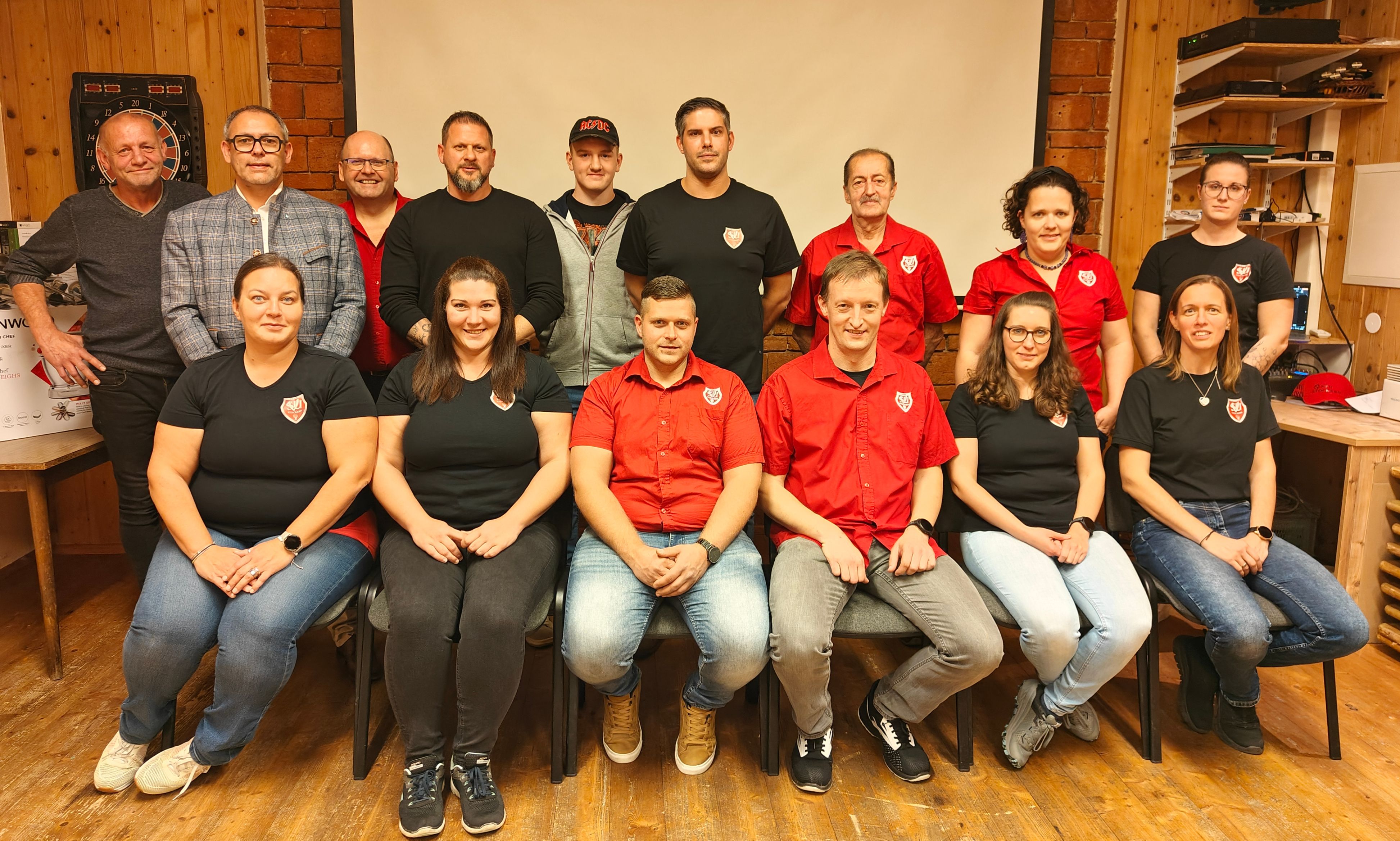 Eine Gruppe von Menschen in roten und schwarzen Shirts und Jeans sitzt auf Stühlen und posiert für ein Foto. Sie stehen vor einer Ziegelwand.