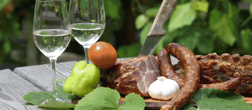 Zwei Weingläser, ein Messer, eine grüne Paprika, eine Knoblauchzehe, Tomaten und verschiedene Würste liegen auf einem Tisch.
